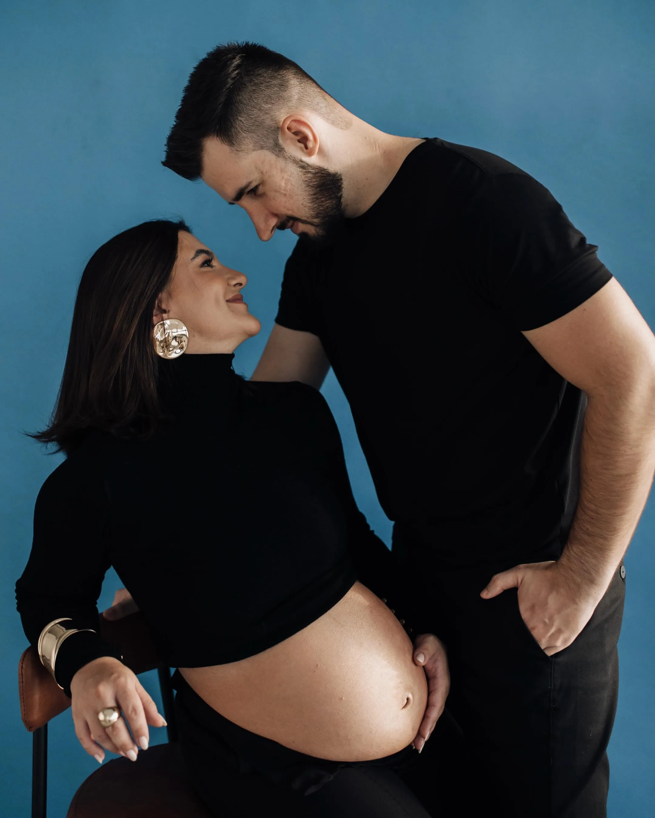 Casal com roupas pretas se olha diante de um fundo azul, com as mãos junto à barriga da mulher