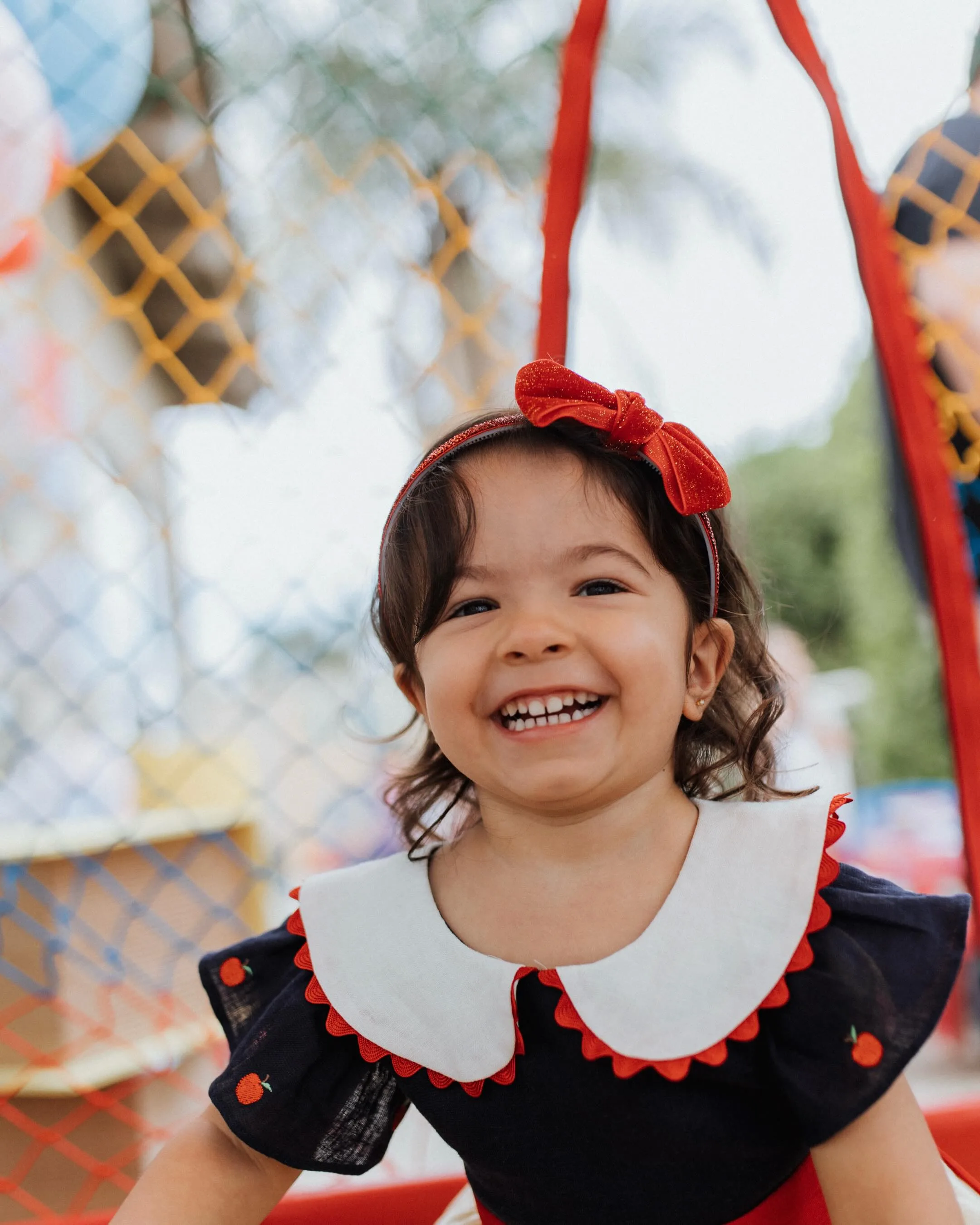 Criança sorridente de laço vermelho posa ao ar livre em um cenário de festa