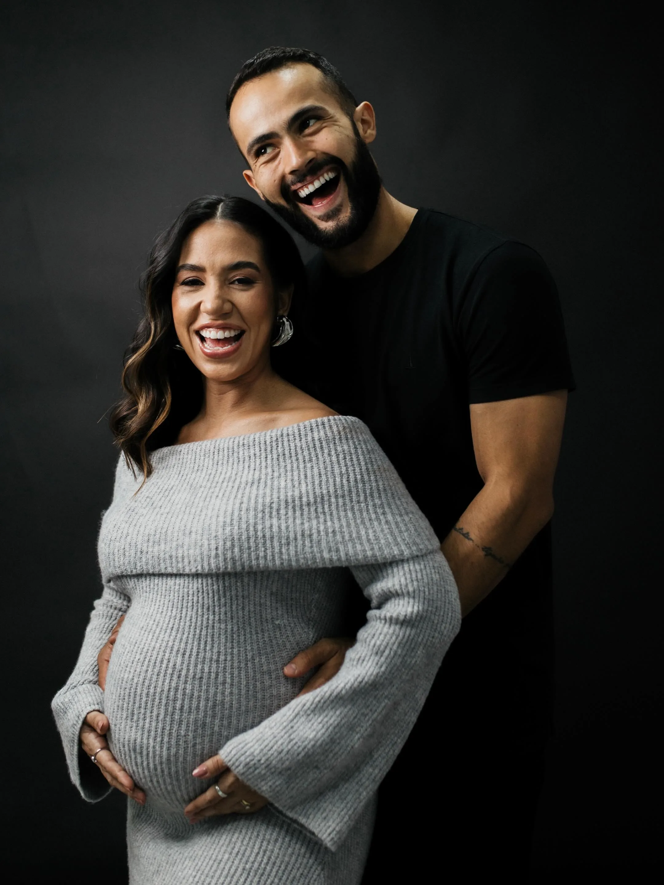 Casal se abraça diante de um fundo escuro, com a mulher usando um vestido cinza